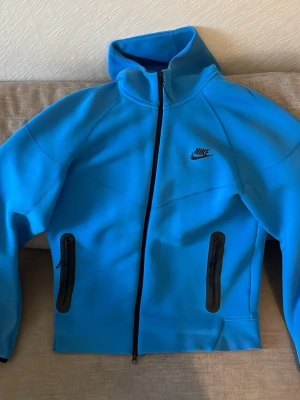Blå zip hoodie från Nike - Snygg blå hoodie från Nike med hel dragkedja framtill och svarta detaljer. Hoodien har två fickor med dragkedja och Nike-logga på bröstet. Tillverkad i ett mjukt och stretchigt material som känns sportigt och modernt. Perfekt för dig som gillar streetwear och sportig stil.