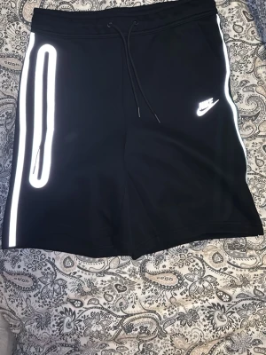 Nike tech reflective shorts - Svarta shorts från Nike med vita reflexdetaljer längs sidorna och logga på vänster ben. Modellen har snörning i midjan och en sportig look. Perfekta för träning eller chill. Tillverkade i mjukt syntetmaterial för skön passform.
