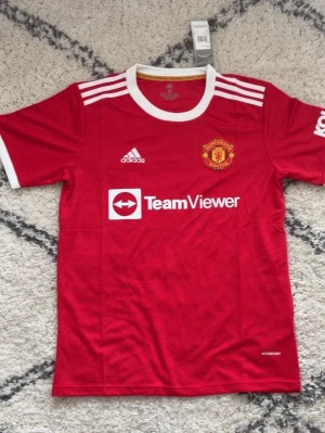 Manchester United matchtröja Adidas - Röd Manchester United matchtröja från Adidas med vita detaljer och klubbmärke på bröstet. Tröjan har korta ärmar, rund halsringning och TeamViewer-logga framtill. Perfekt för fotboll och träning, tillverkad i lätt polyester.