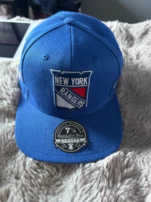 Blå New York Rangers keps - Säljer en blå keps från Mitchell & Ness med New York Rangers broderad logga framtill. Klassisk böjd skärm och snygg sticker kvar på skärmen. Kepsen är tillverkad i ett slitstarkt tyg och har en sportig look som passar perfekt till streetwear.
