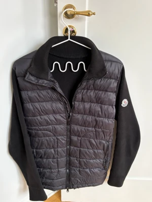 Svart cardiganjacka från Moncler - Snygg svart cardiganjacka från Moncler med quiltad front och stickade ärmar. Jackan har dragkedja framtill, två fickor med dragkedja och en klassisk Moncler-logga på ärmen. Perfekt för lager-på-lager och streetstyle.