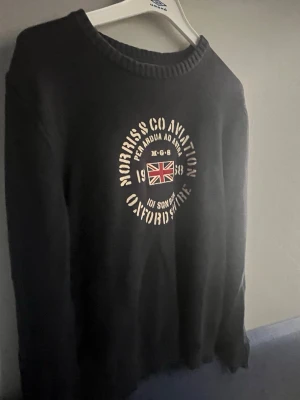 Svart sweatshirt från Morris - Cool svart sweatshirt från Morris med tryck i vitt och Union Jack-flagga på bröstet. Rund halsringning med ribbad kant och avslappnad passform. Perfekt för dig som gillar klassisk brittisk stil och vill ha något bekvämt och snyggt.