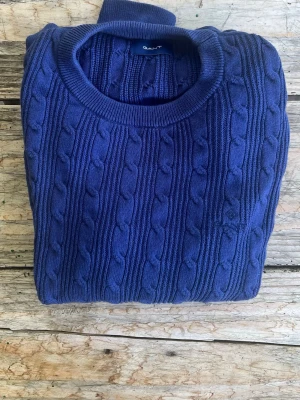 Blå kabelstickad tröja från Gant - Snygg blå kabelstickad tröja från Gant med rund halsringning och diskret broderad logga på bröstet. Tröjan har ett klassiskt mönster och är tillverkad i mjukt stickat material som passar perfekt till alla tillfällen.