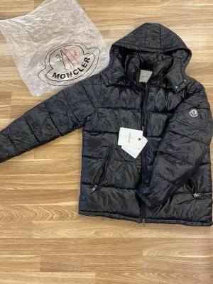 Svart dunjacka från Moncler med huva - Snygg svart dunjacka från Moncler med glansig finish och avtagbar huva. Jackan har två dragkedjefickor fram, Moncler-logga på ärmen och diskreta detaljer. Perfekt för kalla vinterdagar och riktigt trendig look.