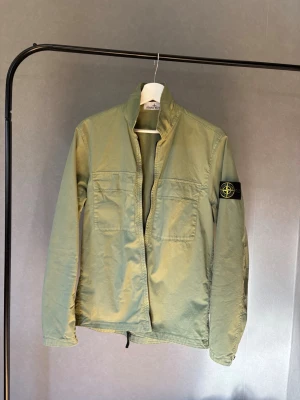 Grön overshirt från Stone Island - Jjusgrön overshirt från Stone Island med patch på vänster ärm. Jackan har två stora bröstfickor, dragkedja framtill och en rak passform. 