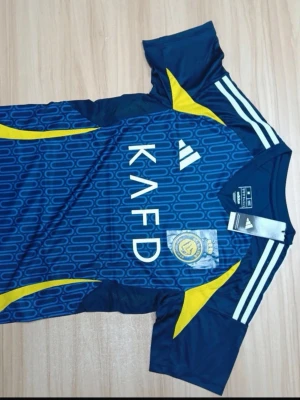 Blå Al Nassr matchtröja från Adidas - Snygg blå matchtröja från Adidas för Al Nassr med gula detaljer och vita ränder på axlarna. Tröjan har klubbmärke och sponsortryck på bröstet samt ett mönster över hela framsidan. Tillverkad i lätt och ventilerande funktionsmaterial, perfekt för träning eller match.