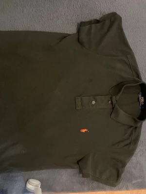 Svart pikétröja från Ralph Lauren - Snygg svart pikétröja från Polo Ralph Lauren med klassisk krage och knappar framtill. Tröjan har korta ärmar och den ikoniska broderade loggan i orange på bröstet. Perfekt för en stilren och avslappnad look.