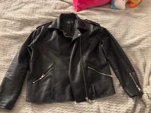 Svart biker skinnjacka från H&M - Snygg svart biker skinnjacka från missguided  med asymmetrisk dragkedja, bred krage och silverfärgade detaljer. Jackan har tre dragkedjeförsedda fickor och axelklaffar med tryckknappar. Tillverkad i konstläder som ger en cool look. Med medföljande skärp 