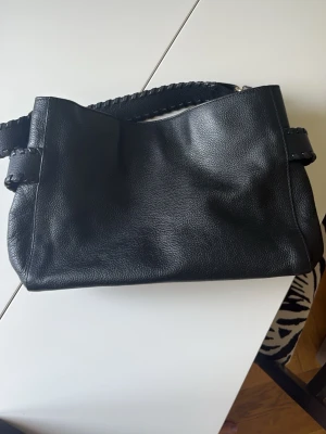 Svart handväska i läderlook - Snygg svart handväska i läderimitation med grovt flätat handtag och guldfärgade metalldetaljer. Väskan har en mjuk, rektangulär form och stängs med dragkedja. 