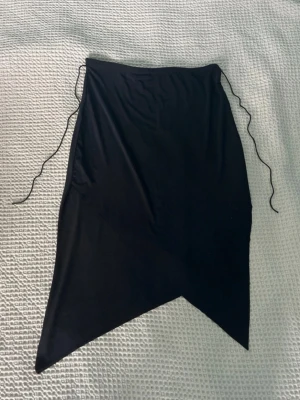 Vintage svart kjol - Trendig svart kjol med snörning i sidorna och asymmetrisk spetsig nederkant. Kjolen har en enkel design och är perfekt för att skapa en edgy look. Tillverkad i mjukt material som ger en bekväm passform.