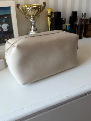 Beige necessär från Burberry - Stilren beige necessär från Burberry i mjukt material med präglad logga på framsidan. Rektangulär form med dragkedja upptill, perfekt för att organisera dina småsaker eller smink. Enkel och snygg design som passar till allt. OBS: dom 5 första bilderna är tagna från en annan, den jag säljer är helt oanvänd och oöppnad! Ställ gärna frågor om storlek medmera! 😉🙂