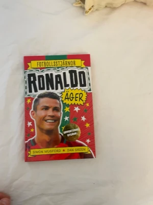 Ronaldo Äger - Fotbollsstjärnor - Färgstark bok om fotbollsstjärnan Ronaldo, skriven av Simon Mugford och Dan Green. Omslaget är rött med gröna och gula detaljer, dekorerat med stjärnor och illustrationer. Perfekt för dig som gillar sport och inspirerande berättelser.