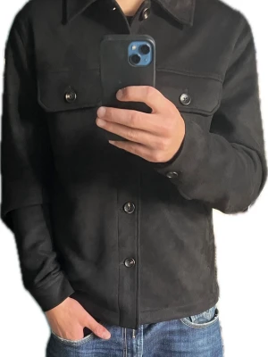 Svart overshirt - Stilren svart overshirt- mockamaterial med klassisk krage och två stora bröstfickor med knappar. Jackan har raka ärmar och knäppning framtill med metallknappar. Passar perfekt till jeans och hoodie för en avslappnad look.