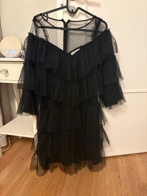 Svart volangklänning från Joelle - Säljer en svart offshoulder klänning från Joelle med flera lager volanger och transparent mesh. Klänningen har prickigt mönster upptill och är perfekt för dig som vill sticka ut. Luftig och trendig design med romantiska detaljer.