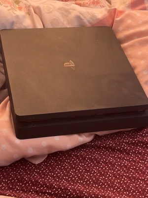 PlayStation 4 Slim svart konsol - Säljer en svart PlayStation 4 Slim från Sony. Konsolen har en stilren, fyrkantig design med rundade hörn och PlayStation-loggan i silver på ovansidan. Perfekt för gaming och streaming hemma.