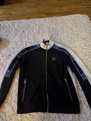Svartvit zip hoodie från EA7 - Snygg svart och vit hoodie från EA7 Emporio Armani med dragkedja framtill och två fickor med dragkedja. EA7-logga på bröstet och blåa detaljer med logotyp längs ärmarna. Tillverkad i mjukt material, perfekt för en sportig och trendig look.