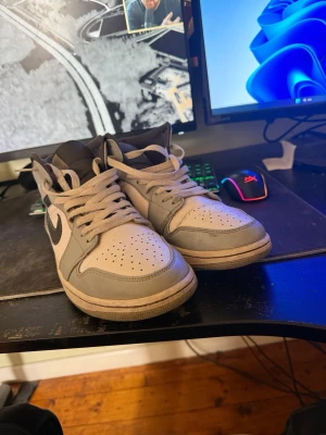 Nike Air Jordan 1 Mid grå/vit - Säljer ett par Nike Air Jordan 1 Mid sneakers i grått och vitt med svarta detaljer. Klassisk siluett med perforerad tåbox, vita skosnören och ikoniska Swoosh-loggan på sidan. Tillverkade i läder och syntetmaterial för en clean och stilren look. Lite slitet på sulan som syns på bilden men känns inget o när man går.