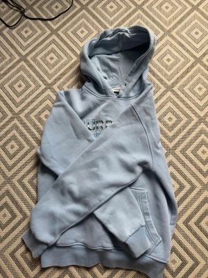 Ljusblå Burberry hoodie med tryck - Säljer en ljusblå hoodie från Burberry med klassiskt tryck på bröstet där det står 'BURBERRY LONDON ENGLAND'. Hoodien har huva och en stor magficka framtill. Perfekt för dig som gillar stilrena och exklusiva plagg med streetkänsla.