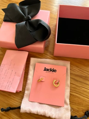 Guldpläterade örhängen från Jackie - Helt nya örhängen från Jackie i guldpläterad metall. De har en halvcirkelformad design med små taggar. Levereras i originalask och tygpåse – perfekt för dig som gillar trendiga accessoarer. Jag har aldrig använt dem, då jag fick dem i julklapp och de inte är i min smak. Kvitto finns. Originalpris 599kr