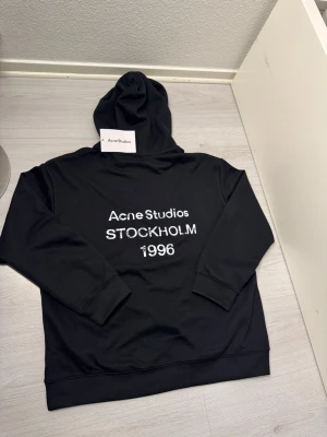 Svart hoodie Acne Studios Stockholm - Snygg svart hoodie från Acne Studios med tryck på ryggen där det står 'Acne Studios STOCKHOLM 1996'. Klassisk passform med huva och ribbade muddar. Tillverkad i mjukt bomullsmaterial som känns skönt mot huden. Perfekt för en clean och stilren look. Ej använd, fraktar samma dag eller dagen efter. Längden 67cm bredden från axel till axel 54 cm . Fler frågor kom DM :)