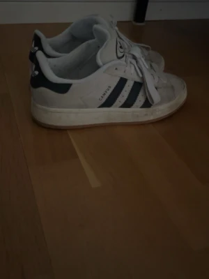 Adidas Campus beige sneakers - Klassiska Adidas Campus sneakers i vitt med svarta ränder och logga på hälen. Skorna har lågt skaft, snörning och är tillverkade i mocka och gummi. Perfekta för en avslappnad streetwear-look. Passar mig som är 38 