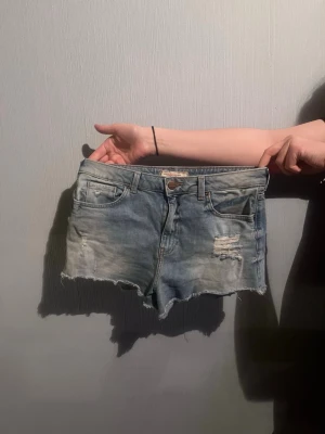 Blå jeansshorts med fransig kant - Säljer ett par ljusblå jeansshorts med fransig kant och slitna detaljer framtill. Klassisk femficksmodell med knapp och dragkedja. Perfekta för sommaren🫶🏻