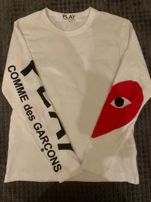 Långärmad Comme des Garçons PLAY - Säljer en vit långärmad t-shirt från Comme des Garçons PLAY med svart tryck längs ena ärmen och det ikoniska röda hjärtat med öga på andra ärmen. Tillverkad i mjuk bomull och har en clean, modern look. Storlek S