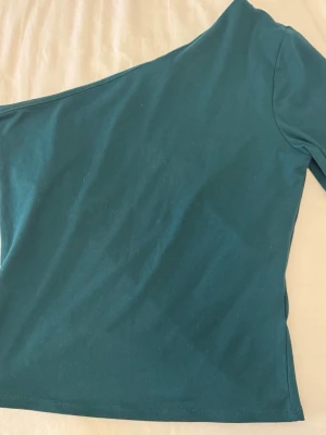 Mörkgrön oneshoulder topp - Snygg mörkgrön oneshoulder topp i mjukt material. Toppen har lång ärm på ena sidan och är axelbandslös på andra, vilket ger en trendig och unik look. Perfekt att styla med jeans eller kjol för en cool outfit.