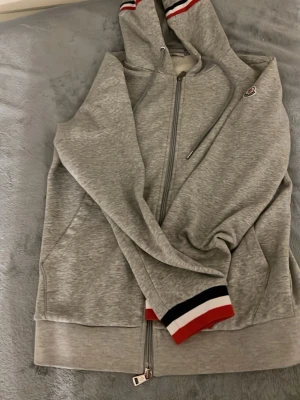 Grå Moncler hoodie med dragkedja - Snygg grå hoodie från Moncler med hel dragkedja, huva och ribbade muddar med röd, vit och svart rand. Klassisk Moncler-logga på bröstet och detaljer i samma färger på huvan. Tillverkad i mjukt bomullsmaterial för skön känsla.