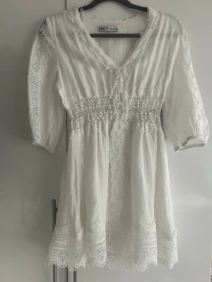 Vit spetsklänning från Zara - Vit kortklänning från Zara med v-ringning, spetsdetaljer längs ärmar, nederkant och fram, samt smock i midjan. Klänningen har trekvartsärm och är tillverkad i ett lätt material som passar perfekt till varma dagar, student, avslutning. 