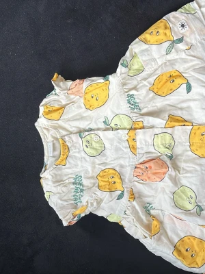 Vit kortklänning med fruktmönster - Vit kortklänning i bomull med färgglatt mönster av citroner, lime och apelsiner i gult, grönt och orange. Klänningen har korta ärmar, rund hals och knappar framtill. Lekfull och somrig design som sticker ut.