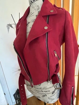 Vinröd mocka jacka med bälte - vinröd mocka jacka i bikerstil med silvriga dragkedjor och knappar. Jackan har bred krage, bälte i midjan och detaljerade axelpartier. Perfekt för dig som vill sticka ut med en cool och trendig look.