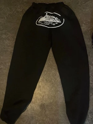 Svarta mjukisbyxor corteiz - Säljer ett par svarta mjukisbyxor med resår i midjan och ett stort vitt tryck av en byggnad på baksidan. Byxorna har avslappnad passform och är perfekta för chill dagar. Materialet känns mjukt och bekvämt, troligen bomull eller bomullsblandning.