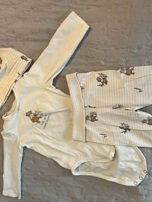 Sött set med björnmotiv för baby - Tre delar: långärmad body, byxor och mössa i mjuk, ljusbeige bomull med söta björnar och växtmotiv. Bodyn har tryckknappar i grenen och byxorna har bred, ribbad midja. Perfekt för mysiga dagar hemma.
