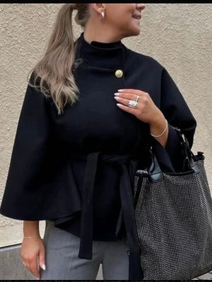 Svart cape-kappa med knytband - Stilren svart poncho-kappa från missäy med hög krage, dekorativ guldfärgad knapp och knytband i midjan. Kappan har vida ärmar och en elegant, minimalistisk design som passar perfekt till höst och vår. Storlek är onesize 