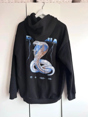 Svart hoodie  med ormtryck. S.  - Snygg hoodie från Black Squad. Svart bottenfärg. Stort tryck på ryggen. Storlek S. Lite ytludd. Ej blekt. Trycket helt o fint. Den är i fint skick.  