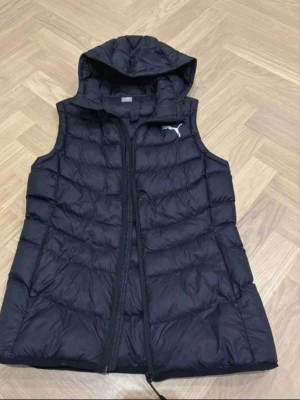 Svart dunväst från Puma med huva - Snygg svart dunväst från Puma med huva och dragkedja framtill. Västen har ett quiltat mönster och den klassiska Puma-loggan på bröstet. Perfekt för lager på lager och streetwear-stil. Materialet är lätt och värmande.