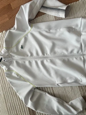 Vit ziptröja från Lacoste - Snygg vit ziptröja från Lacoste med diskreta limegröna sömmar och svarta rutiga detaljer längs ärmarna. Tröjan har hög krage, dragkedja framtill och två fickor. Klassisk Lacoste-logga på bröstet och tryckt logga i nacken.