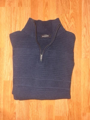 Mörkblå stickad tröja Dressmann - Mörkblå stickad tröja från Dressmann med halv dragkedja och hög krage. Tröjan har diskreta horisontella ränder i stickningen och långa ärmar. Perfekt för lager på lager och en stilren look.