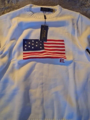 Ralph lauren USA flag  - Vit stickad tröja från Polo Ralph Lauren med amerikansk flagga broderad på bröstet och RL-detalj. Tröjan har rund halsringning och ribbade muddar. Perfekt för dig som gillar klassisk och stilren design.