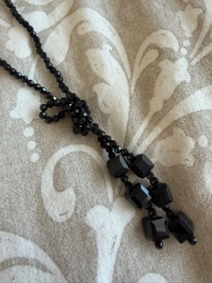Svart halsband med glaspärlor - Snyggt svart halsband med små glaspärlor och dekorativa, större svarta pärlor i geometriska former. Halsbandet har en längre modell och en knutdetalj framtill som ger en cool touch. Perfekt accessoar för att lyfta din outfit med en elegant och mystisk vibe.