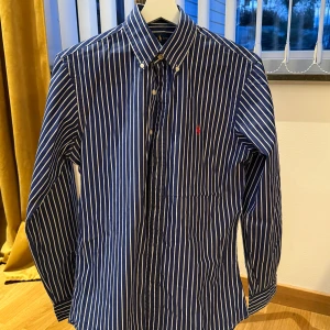 Blårandig skjorta från Ralph Lauren - Klassisk blårandig skjorta från Ralph Lauren med vit knappslå och button-down krage. Skjortan har lång ärm och en liten röd broderad logga på bröstet. Tillverkad i bomull och har en tidlös, normal passform.