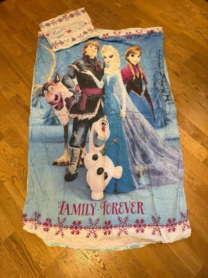 Frost påslakanset Family Forever - Säljer ett påslakanset med motiv från Disney-filmen Frost. Setet är blått och vitt med karaktärerna Elsa, Anna, Olaf och Sven samt texten 'Family Forever' och lila snöflingor. Perfekt för dig som älskar Frost och vill ha mysiga sovstunder.