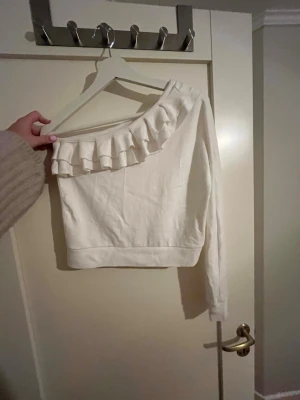 Volang topp - Säljer en vit oneshoulder topp med lång ärm och snygg volangdetalj längs halsringningen. Toppen är croppad och har en modern, stilren look. 