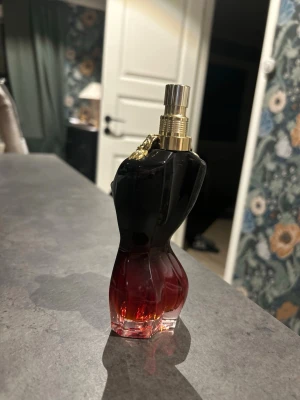 Jean Paul Gaultier La Belle Le Parfum - En elegant parfymflaska från Jean Paul Gaultier formad som en kvinnokropp. Flaskan har en snygg färgskiftning från mörk svart upptill till röd nedtill och är dekorerad med guldfärgade rosor runt halsen. Perfekt för dig som gillar lyxiga och unika dofter.