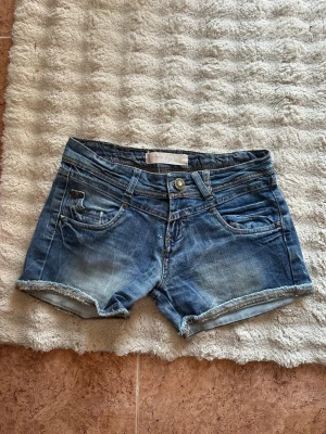 Blå jeansshorts med rå kant - Snygga blå jeansshorts med rå kant och slitningar. Klassisk femficksmodell med knapp och dragkedja framtill samt dekorativa bakfickor med knappar. Perfekta för sommaren och har en avslappnad, casual vibe.