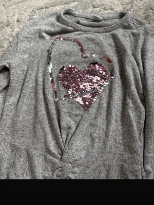 Grå långärmad topp med paljett-hjärta - Grå långärmad topp från Lindex med ett stort hjärta i rosa och silver paljetter på bröstet. Toppen har en twistad detalj framtill och är gjord i mjukt material, perfekt för en avslappnad look.