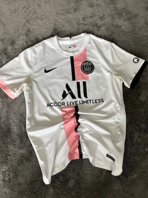 PSG vit matchtröja  - Snygg vit PSG matchtröja med rosa och svart rand längs framsidan. Tröjan har klubbmärke på bröstet och sponsortryck. Tillverkad i lätt och bekvämt material, perfekt för fotboll eller träning.