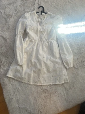 Vit kort klänning med broderade blommor - Söt vit kortklänning med broderade blommor och långa puffärmar. Klänningen har knytband vid halsen och är tillverkad i ett lätt bomullstyg. Perfekt för sommarens alla tillfällen!