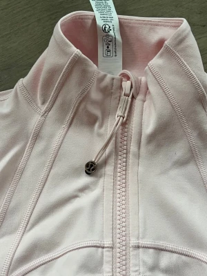 Ljusrosa zipjacka från Lululemon - Sportig ljusrosa zipjacka från Lululemon med hög krage, långa ärmar och figurnära passform. Jackan har dragkedja framtill, snörning vid halsen och två fickor med dragkedja. Perfekt för en aktiv vardag eller träning.